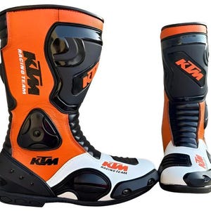 Puede incluir: Par de botas de moto KTM Racing Team naranjas, negras y blancas. Las botas son de caña alta con paneles protectores y el logotipo de KTM. Diseñadas para carreras, son robustas.