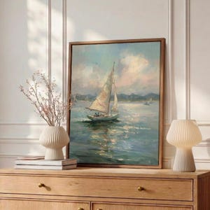 May include: A framed oil painting depicting a sailboat on calm water under a soft, cloudy sky. The artwork features muted blue, white, and pale pink tones, capturing the reflection of the boat on the water surface in a realistic, painterly style.