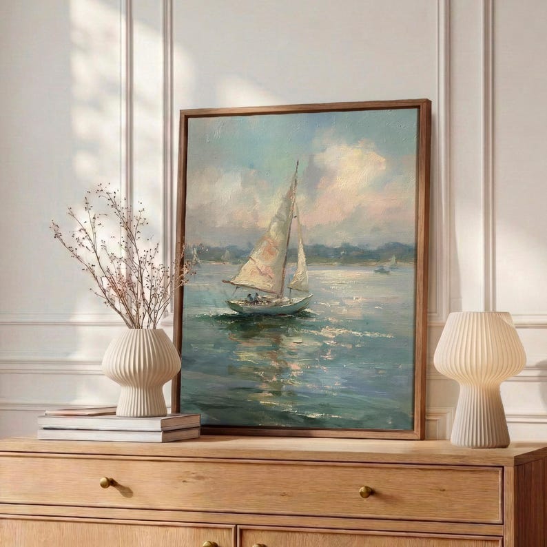 May include: A framed oil painting depicting a sailboat on calm water under a soft, cloudy sky. The artwork features muted blue, white, and pale pink tones, capturing the reflection of the boat on the water surface in a realistic, painterly style.