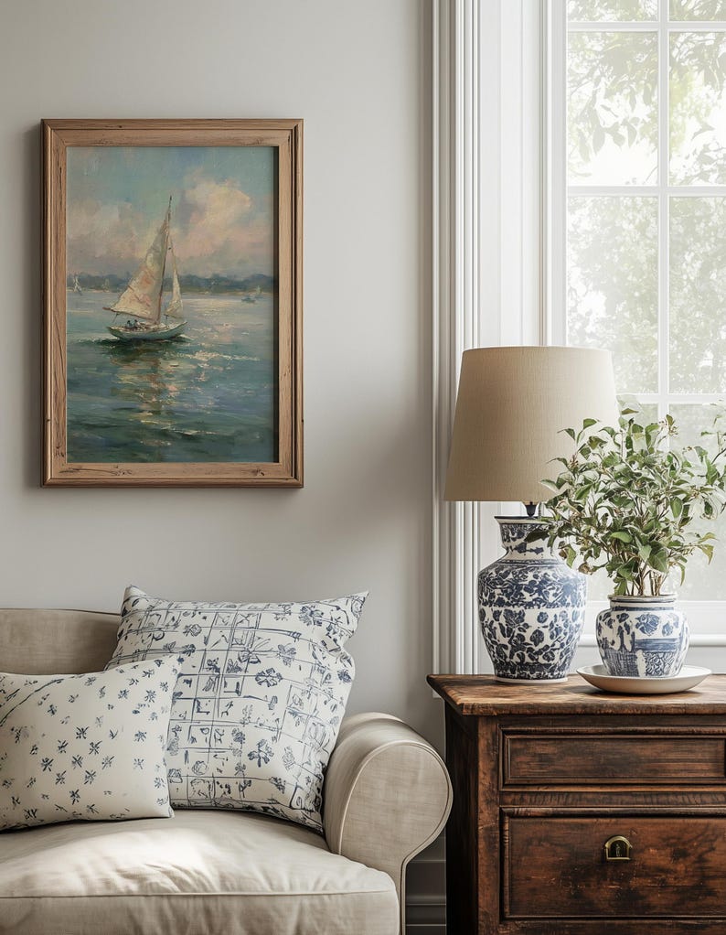 May include: A framed oil painting of a sailboat on water, a beige sofa with two patterned throw pillows, a wooden side table, a blue and white ceramic lamp with a beige shade, and a blue and white ceramic planter with a green plant.