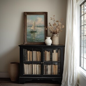 May include: A black wooden cabinet with glass doors containing rows of books. On top sits a framed oil painting of a sailboat on water, a small white ceramic vase, and a larger tan ceramic vase filled with dried branches of brown foliage.