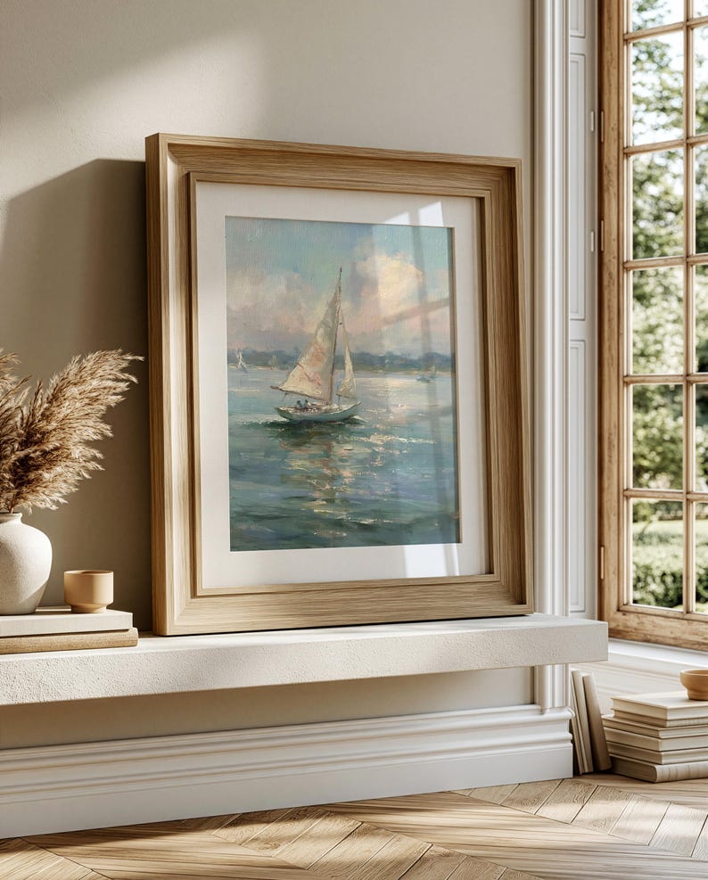 May include: A framed painting of a sailboat on calm water under a soft, cloudy sky. The artwork features muted blue and white tones, depicting a single vessel with sails raised. It is encased in a light-toned wooden frame with a white mat border.