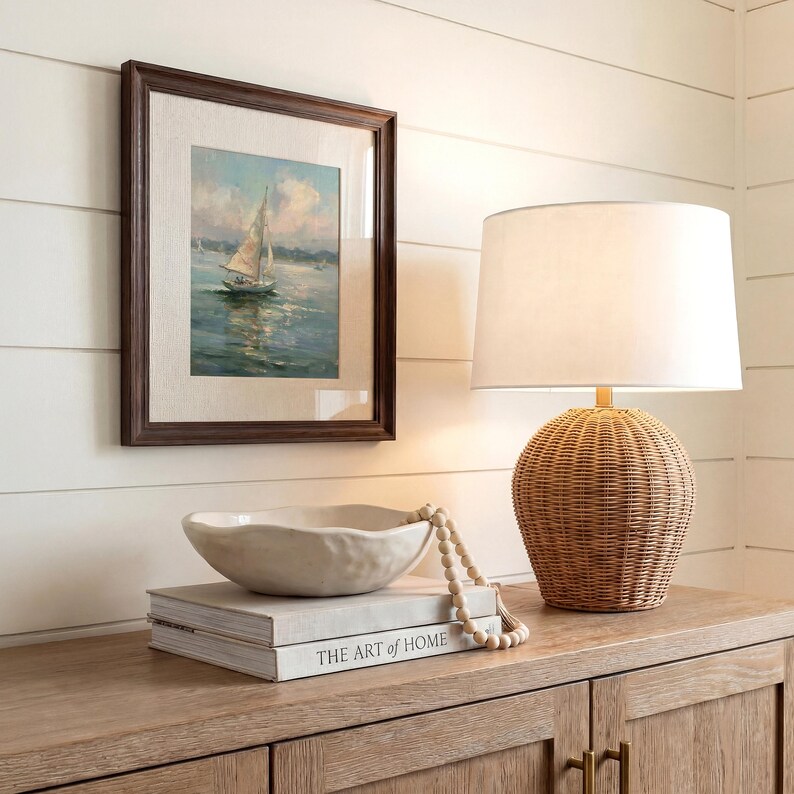 May include: A framed painting of a sailboat on water, a white lamp with a woven rattan base, and a cream-colored ceramic bowl resting on a stack of books titled The Art of Home. The scene features a neutral color palette with natural textures.