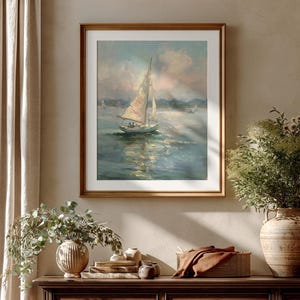 May include: A framed oil painting depicting a sailboat with white sails on calm, reflective water under a soft, cloudy sky. The artwork features muted blue, white, and tan tones, presented in a simple wooden frame with a white mat border.