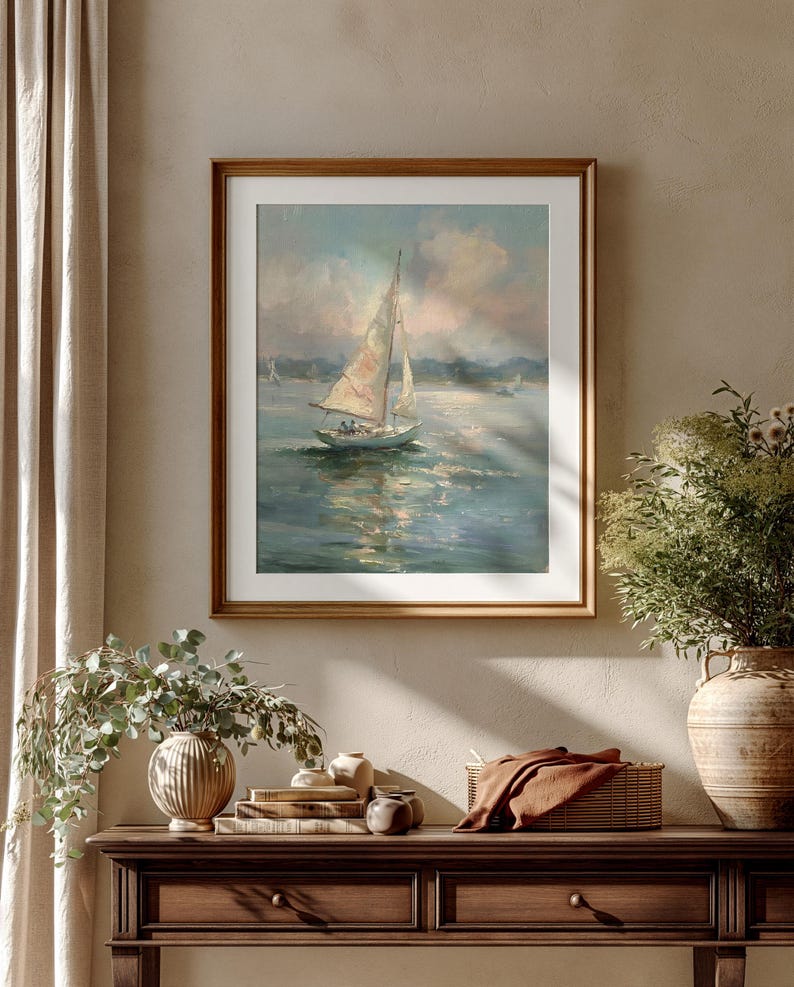 May include: A framed oil painting depicting a sailboat with white sails on calm, reflective water under a soft, cloudy sky. The artwork features muted blue, white, and tan tones, presented in a simple wooden frame with a white mat border.