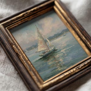 May include: An oil painting of a sailboat on water, framed in a dark brown and gold ornate wooden frame. The artwork depicts a white sailboat with sails catching the wind, set against a soft, muted sky and textured blue-green water surface.