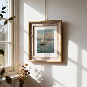 May include: A framed painting of a sailboat on water under a cloudy sky, enclosed in a rustic, weathered light brown wooden frame. The artwork is matted with a white border and hangs from a thin string against a plain white wall surface.