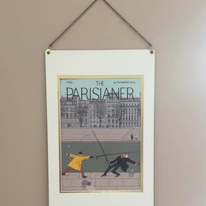 Può includere: Stampa incorniciata della copertina della rivista "The Parisianer", con una scena a fumetti di due uomini che pescano sulla riva di un fiume. L'opera d'arte ha un bordo dorato ed è sospesa da una catena. Lo sfondo è una parete marrone chiaro.