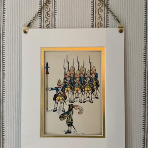 May include: Framed print depicting a line of soldiers in blue coats and yellow pants, with a drummer in the foreground. The print is in a white frame with a gold border, hanging from a chain.