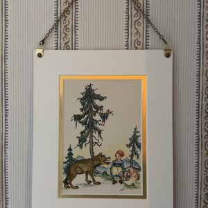 May include: Framed vintage illustration depicting a wolf and a child in a forest setting. The artwork features a white frame with a gold border, suspended by a chain. The background is a striped wallpaper.
