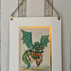 May include: Framed illustration featuring a green dragon carrying soldiers with rifles. The artwork has a gold border and is set in a white frame, suspended by a chain. The background is a striped wallpaper.