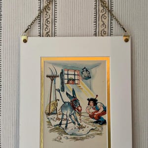 May include: A framed illustration depicting a donkey surrounded by coins, with a man and a window in the background. The artwork is in a white frame with a gold border, suspended by a chain.