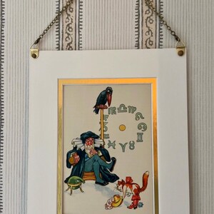 May include: A framed vintage print featuring a seated figure with a long beard, a raven, and zodiac symbols. The artwork is in a white frame with a gold border, suspended by a chain. The print has a whimsical, antique aesthetic.