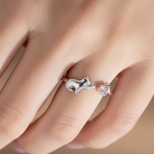 Puede incluir: Un anillo plateado con un diseño de gato que se extiende hacia una gema rosa en forma de corazón. El anillo se lleva en un dedo, mostrando los delicados detalles del gato y el corazón. Es una pieza de joyería.
