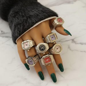 May include: A hand displaying several silver-toned watch rings with various colored faces, including pink, purple, red, and black. The rings have a classic watch design with watch bands and watch faces. The hand has green nail polish.