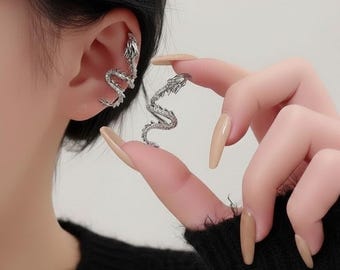 May include: A silver-colored dragon ear cuff and earring set. The ear cuff wraps around the ear, while the earring is held in the fingers. The dragon design features intricate scales and a curved body, creating a unique and stylish accessory.