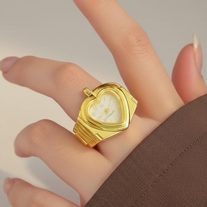 Peut inclure: Une montre-bague dorée avec un cadran en forme de cœur. Le cadran de la montre est blanc avec des chiffres et des aiguilles noirs. La bague a un bracelet rectangulaire et une petite couronne.