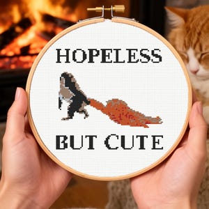 May include: A cross-stitch hoop with the words "HOPELESS BUT CUTE" in black block letters. The design features a black and brown creature reaching for an orange shape. The hoop is held in front of a fireplace and a cat.