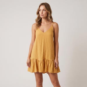 Könnte beinhalten: Ein senfgelbes ärmelloses Minikleid mit V-Ausschnitt, dünnen Spaghettiträgern und einer strukturierten, knittrigen Stoffoberfläche. Das Kleid hat eine lockere A-Linien-Silhouette mit einem gerafften Volantsaum, der über dem Knie endet.