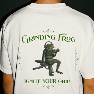 Può includere: T-shirt bianca con grafica verde di una rana che solleva pesi con le cuffie. Il testo "Grinding Frog" e "Ignite Your Chill" sono stampati in verde.