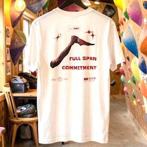 Pode incluir: Camiseta branca com um gráfico de um braço e as palavras "FULL SPAN COMMITMENT" em vermelho. A camiseta está pendurada em um cabide preto. O texto "LIVE BOLDLY" está no topo. O fundo mostra uma parede de escalada.