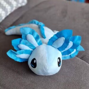Puede incluir: Un peluche de ajolote en tonos azules y blancos. El peluche tiene ojos grandes y expresivos y branquias plumosas. El juguete est&aacute; sobre una superficie de color neutro, mostrando su aspecto suave y tierno. Una criatura caprichosa y encantadora.