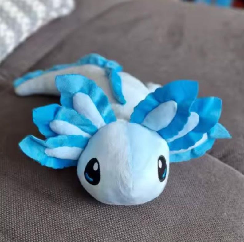 Puede incluir: Un peluche de ajolote en tonos azules y blancos. El peluche tiene ojos grandes y expresivos y branquias plumosas. El juguete est&aacute; sobre una superficie de color neutro, mostrando su aspecto suave y tierno. Una criatura caprichosa y encantadora.