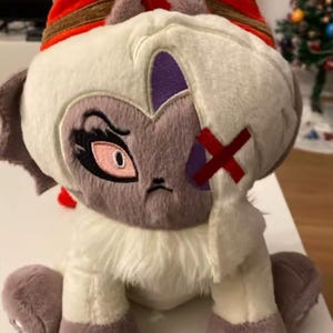 May include: A plush toy featuring a stylized creature with a white body, purple accents, and a red 'X' on its face. The creature has large eyes, a grumpy expression, and striped horns. The toy is soft and cuddly.