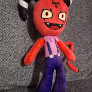 May include: A plush doll featuring a red devil-like character with black and white horns, red arms, and a tail. The doll has a pink shirt, purple pants, and a smiling face with yellow eyes.