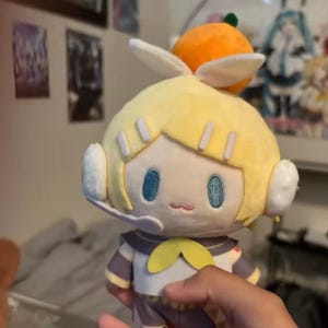 May include: A plush doll of a character with yellow hair, blue eyes, and a small orange fruit on top of its head. The doll is wearing a gray and white outfit with a yellow bow.