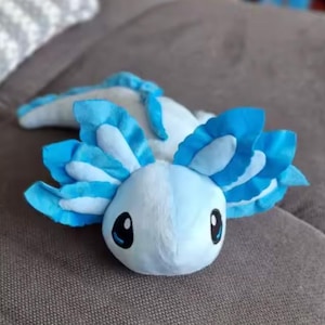 Puede incluir: Un peluche de ajolote en tonos azules y blancos. El peluche tiene ojos grandes y expresivos y branquias plumosas. El peluche est&aacute; sobre una superficie de color neutro.