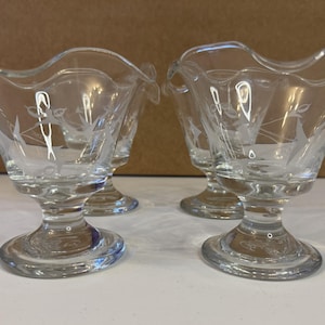 May include: Four clear glass dessert dishes with a decorative etched sailboat design. Each dish has a flared rim and sits on a pedestal base. The dishes are arranged on a white surface.