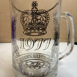 May include: Clear glass mug with a handle, featuring a crown design and the year 1977. The text "The Queen's Silver Jubilee" is also displayed. The mug has a silver rim and decorative elements.