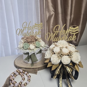 May include: Two floral arrangements in white hat boxes with gold glitter toppers that read "Hajj Mubarak." One arrangement has white, green, and tan flowers. The other has white flowers. A beaded prayer bracelet is in the foreground.