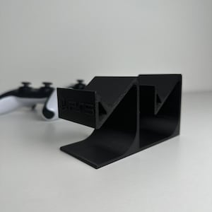 Könnte beinhalten: Ein schwarzer Gaming-Controller-Ständer aus Kunststoff mit strukturierter Oberfläche und V-förmigem Design. Auf der Vorderseite des Ständers ist der Text PS5 eingeprägt. Der Ständer ist so konzipiert, dass er einen Gaming-Controller sicher hält.