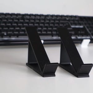 Könnte beinhalten: Zwei schwarze, abgewinkelte Ständer für ein Tablet oder Telefon, mit einer schwarzen Tastatur im Hintergrund. Die Ständer bestehen aus einem soliden Material und haben ein schlichtes, modernes Design. Sie sind so konzipiert, dass sie ein Gerät in einem Winkel halten.