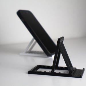 Könnte beinhalten: Ein schwarzer Smartphone-Ständer steht im Fokus, mit einem zweiten, ähnlichen Ständer im Hintergrund. Der Ständer ist so konzipiert, dass er ein Telefon in einem Winkel hält. Der Ständer besteht aus Kunststoff und hat eine rechteckige Basis.