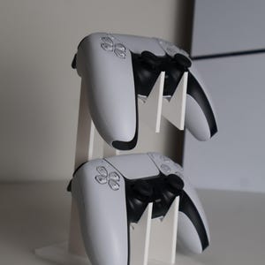 May include: A white gaming controller stand holding two white and black controllers. The stand is made of white material and has a vertical design. The controllers have black accents and button details.