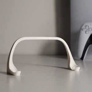 Könnte beinhalten: Ein weißer, gebogener Ständer zur Aufnahme einer Spielkonsole. Der Ständer hat eine glatte, matte Oberfläche und ein minimalistisches Design. Ein weißer Gamecontroller ist im Hintergrund zu sehen.