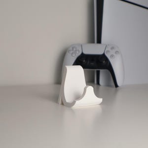 Könnte beinhalten: Ein weißer, minimalistischer Ständer für einen Spielekonsolen-Controller. Der Ständer befindet sich im Vordergrund, mit einem weißen Controller und einer Konsole im Hintergrund. Der Ständer hat ein geschwungenes Design.