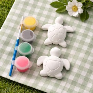 Può includere: Un kit per l'artigianato con due figurine di tartarughe in ceramica bianca, vasetti di vernice gialla, grigia, verde e rosa e un pennello con manico blu. Gli oggetti sono disposti su un tessuto a scacchi verde e bianco, con un fiore bianco sullo sfondo.