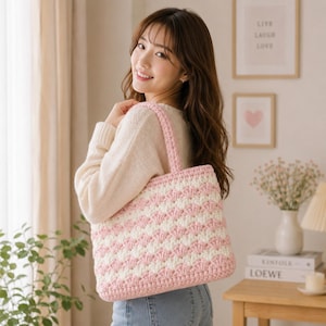 May include: A handmade crochet tote bag in a pink and white striped pattern. The bag has a rectangular shape with a solid pink border and matching handles. The bag is held by a person.