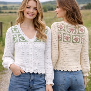 May include: Two women wearing crocheted cardigans. One cardigan is white with green and white floral accents, a scalloped hem, and pearl buttons. The other is cream-colored with pink and green floral accents. Both are paired with blue jeans.