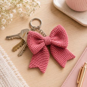 May include: A dusty rose crochet bow keychain featuring a textured, knotted yarn construction. The bow is attached to a silver-toned metal split ring and chain, which holds three standard metal keys. The design is symmetrical with two loops.
