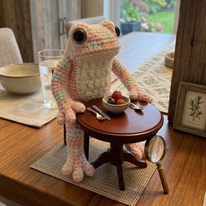 Könnte beinhalten: Eine handgefertigte Häkelfroschpuppe in rosa und blauen Streifen, die an einem kleinen runden Tisch sitzt. Der Frosch hat große schwarze Augen und ist mit einer kleinen Schüssel mit Beeren, einem Löffel und einer Gabel positioniert. Eine Lupe liegt in der Nähe.