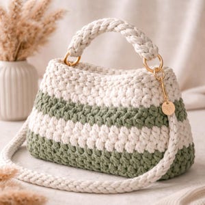 May include: A handmade crochet handbag in cream and olive green stripes. The bag features a braided handle and a detachable shoulder strap. Gold-colored rings attach the handle and strap to the bag. A small gold charm hangs from the side.