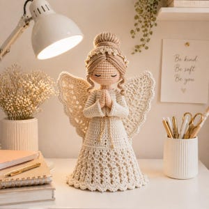 May include: A handcrafted crochet angel doll in cream and beige tones. The doll has closed eyes, a halo, and folded hands. The angel wears a long dress and has detailed wings. The image includes a desk lamp, notebooks, and a sign that says "Be kind, Be soft, Be you."
