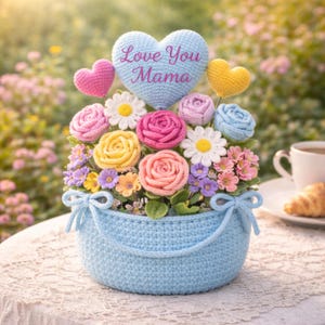 Pode incluir: Uma cesta azul de crochê transbordando de flores coloridas de fio, incluindo rosas e margaridas. Um coração azul com "Love You Mama" está centralizado acima do arranjo, com corações rosa e amarelos menores. Um presente artesanal charmoso.