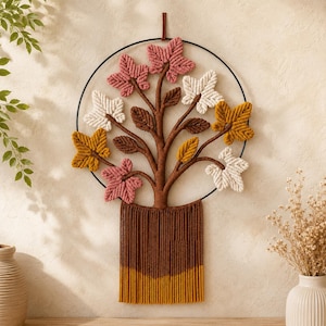 May include: A macrame wall hanging featuring a circular black metal frame containing a tree design with brown cord branches and leaves in shades of pink, cream, and mustard yellow. The base of the tree has a long, straight brown and yellow fringe.