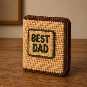 May include: A crocheted rectangular item with the words "BEST DAD" in dark green letters on a tan background. The item has a brown border and is sitting on a wooden surface. The item is a handmade gift.